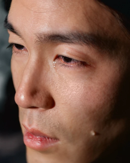 Close-up portrait of an Asian adult male with tears in his eyes, set against a dark, blurred background. Focus on skin texture and details.