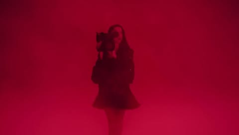 A woman stands illuminated in a red environment, holding a camera and dressed in dark clothing, creating a mysterious and striking image.