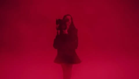A woman stands illuminated in a red environment, holding a camera and dressed in dark clothing, creating a mysterious and striking image.