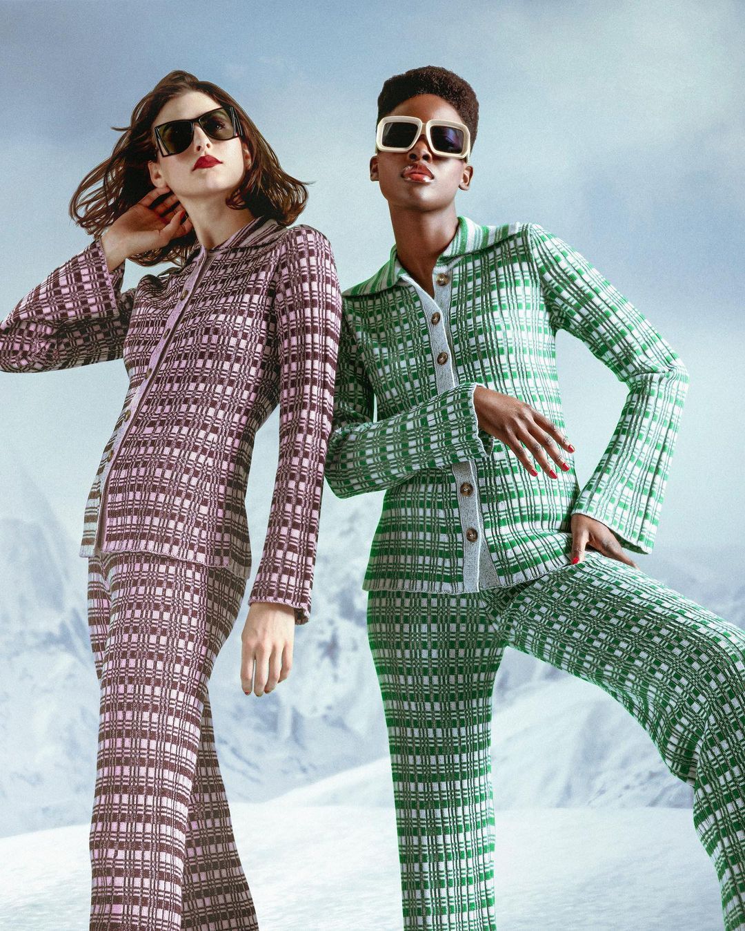 Two diverse women pose in matching patterned knitwear suits and sunglasses in a snowy landscape, showcasing fashion and style.