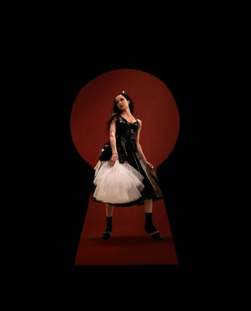 A woman in a black dress with a white tulle skirt poses against a red backdrop, framed by a black keyhole shape, creating a dramatic effect.