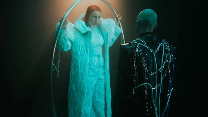 Two people in a dimly lit studio: one wearing a bright quilted coat, the other a black jacket with silver chain details.