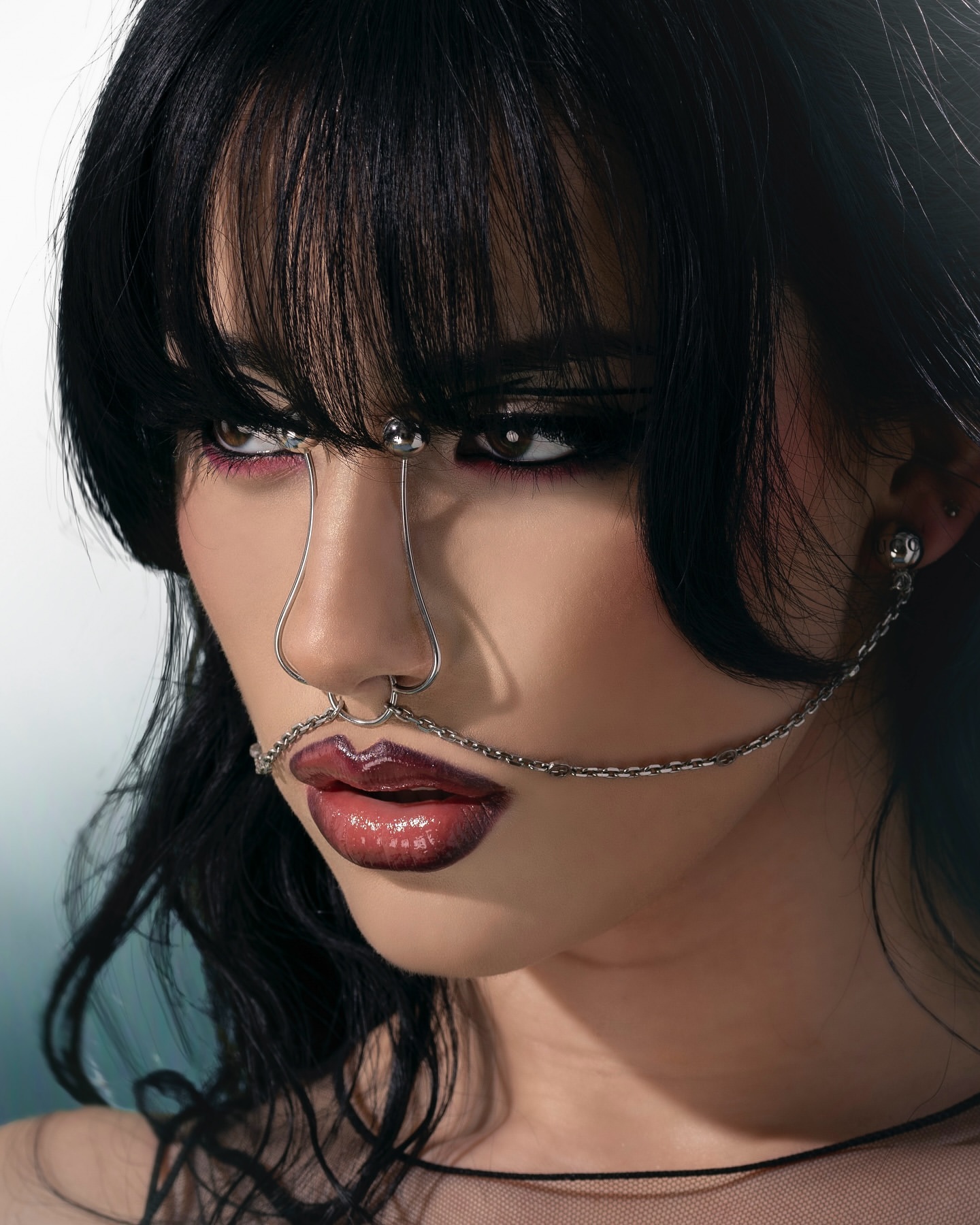 Close-up studio portrait of a female with a jeweled nose chain, dark wavy hair, and glossy burgundy lips against a hazy blue backdrop.