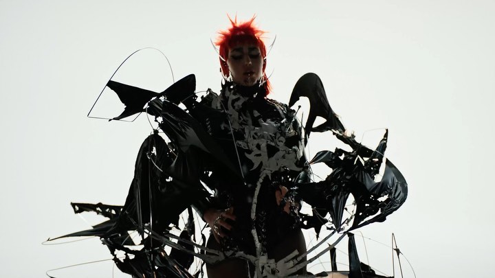 Adult female with bright red hair poses in a dramatic black and white patterned sculptural costume against a stark white backdrop.