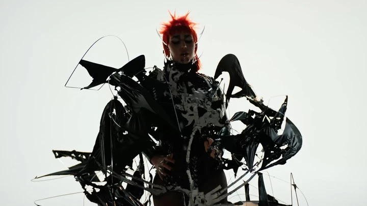 Adult female with bright red hair poses in a dramatic black and white patterned sculptural costume against a stark white backdrop.