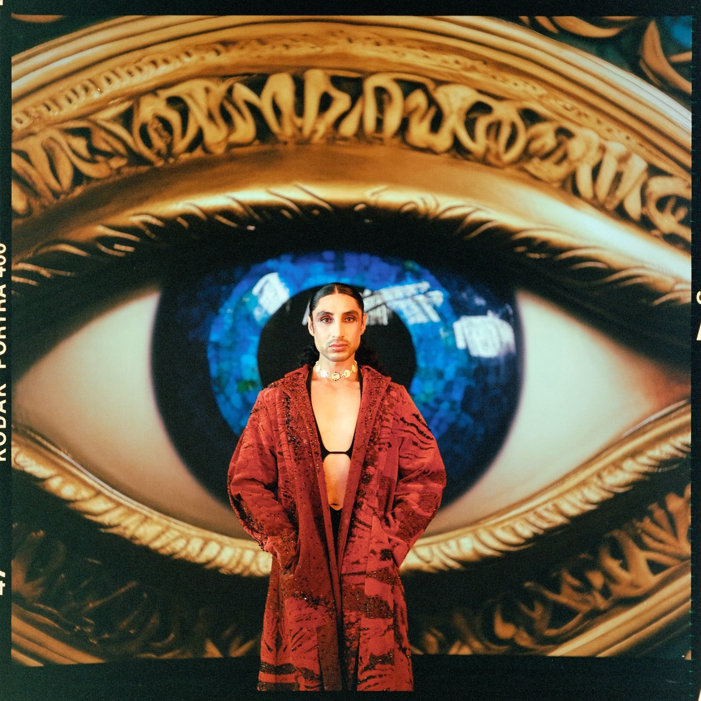Portrait of a man in a red patterned coat standing before a giant illustrated eye backdrop by photographer Sam Cannon.