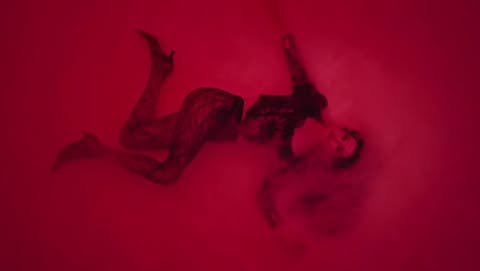 An adult female fashion model poses in a surreal, dramatic studio shot. The woman is laying down, wearing heels, against a solid red backdrop.