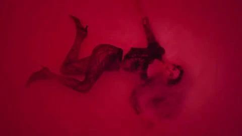 An adult female fashion model poses in a surreal, dramatic studio shot. The woman is laying down, wearing heels, against a solid red backdrop.