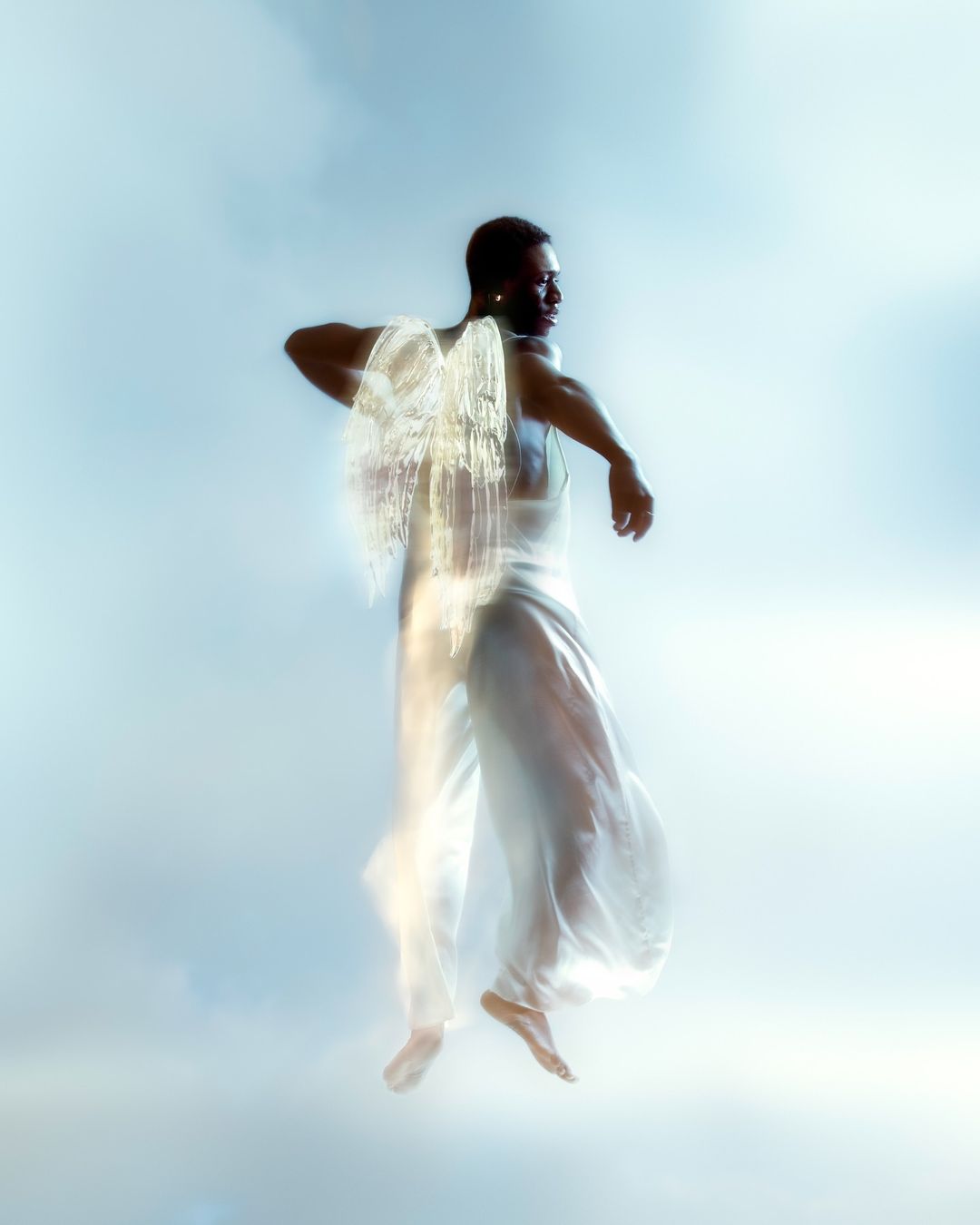African-American man wearing a white outfit with artistic light effects creating faux wings, soft light, against a sky-blue background.