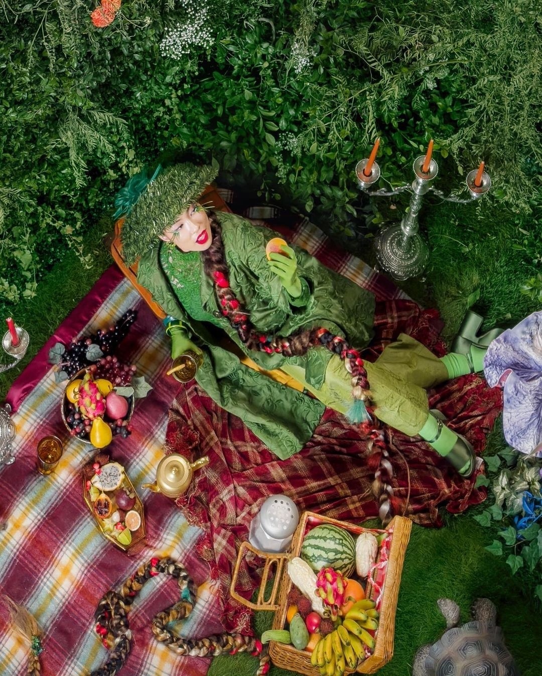 Overhead view of a person in a green costume holding fruit at an outdoor picnic with fruit basket, blanket, candelabra, and foliage.