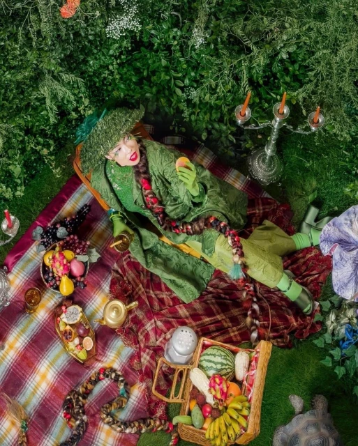 Overhead view of a person in a green costume holding fruit at an outdoor picnic with fruit basket, blanket, candelabra, and foliage.