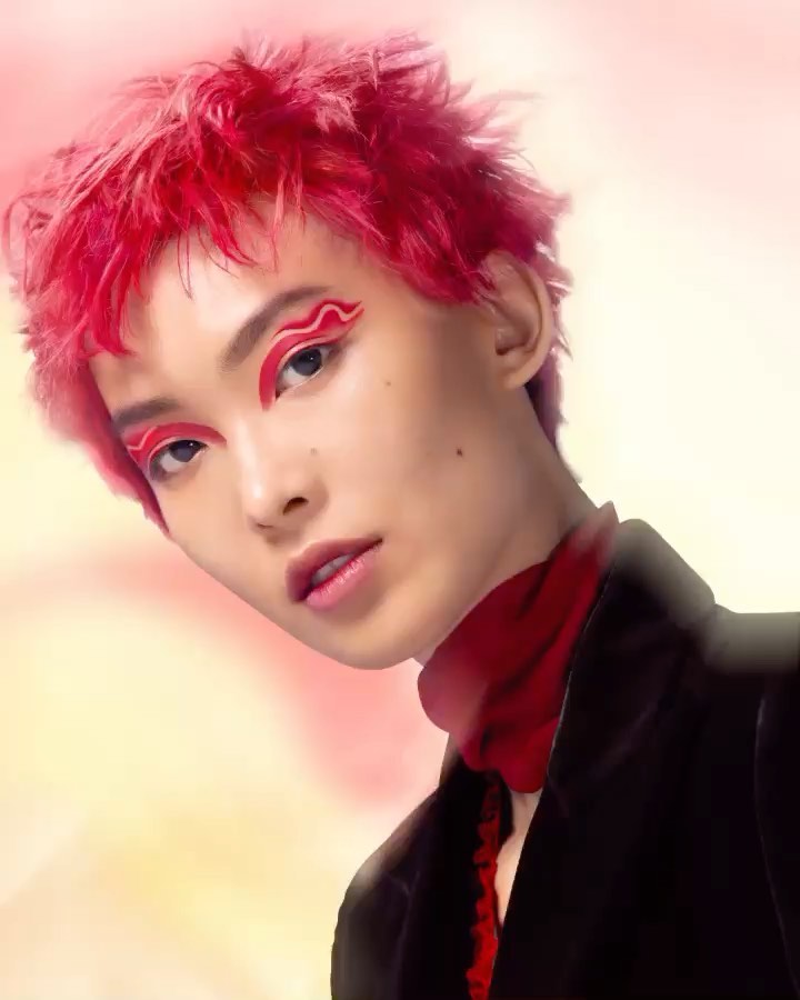 Close-up portrait of a person with bright red hair and dramatic red eye makeup, wearing a black jacket and red scarf on a painterly background.