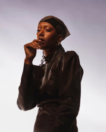 Portrait of a black woman wearing a patterned headscarf and dark top against a light background by Sam Cannon