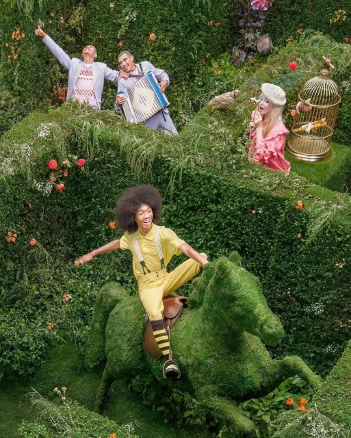A group of people are playfully positioned among lush greenery, one riding a topiary horse, creating a whimsical, surreal, garden scene.