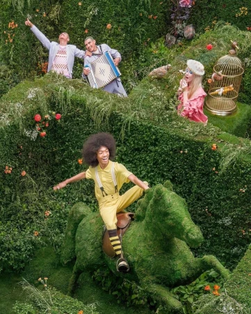 A group of people are playfully positioned among lush greenery, one riding a topiary horse, creating a whimsical, surreal, garden scene.