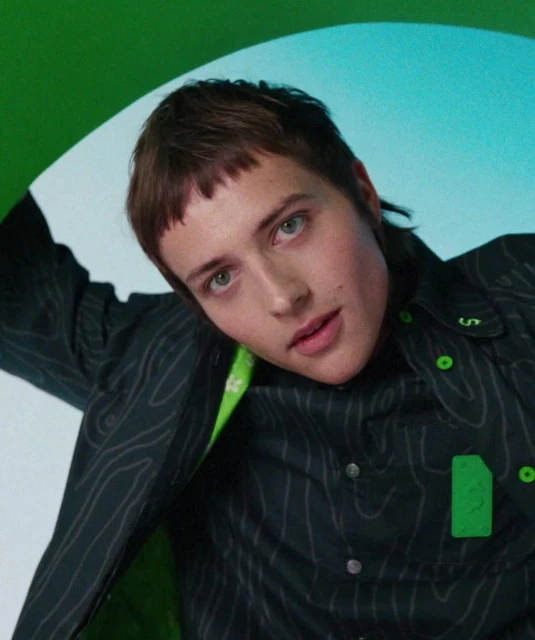 Adult posing in a black button-down shirt with green accents, under green and blue studio lighting, looking directly at the camera.