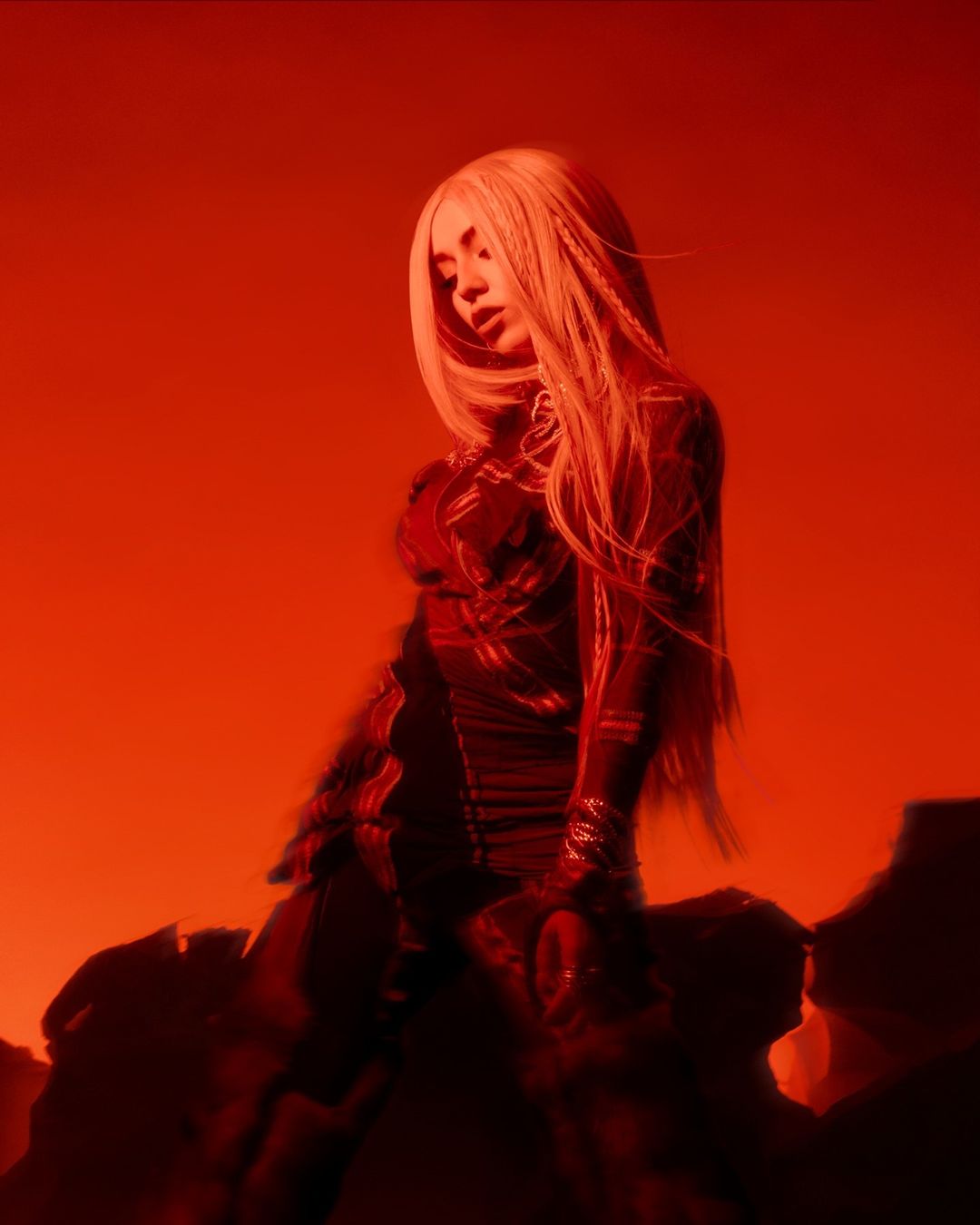 Portrait of a woman with long blonde hair posing in a dark red environment. The model is wearing dark clothing and has a solemn expression.
