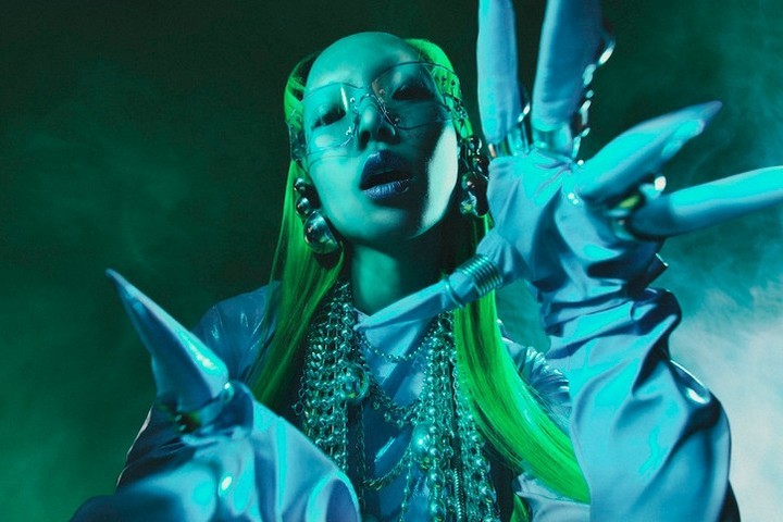Person with green hair, futuristic glasses, sculptural gloves and jewelry in a studio shot with blue and green lighting and fog.