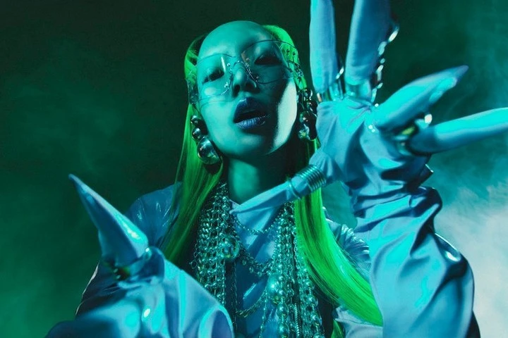 Person with green hair, futuristic glasses, sculptural gloves and jewelry in a studio shot with blue and green lighting and fog.
