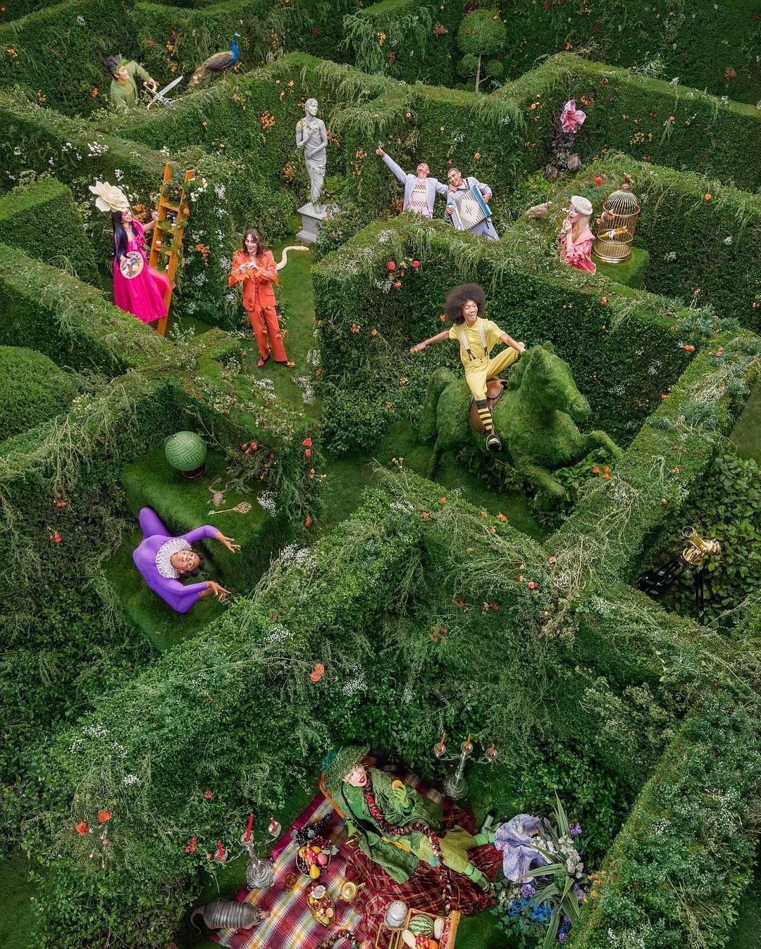 Aerial view of people posing in a garden maze featuring green hedges, topiary art, and whimsical costumes for a surreal fashion shoot.