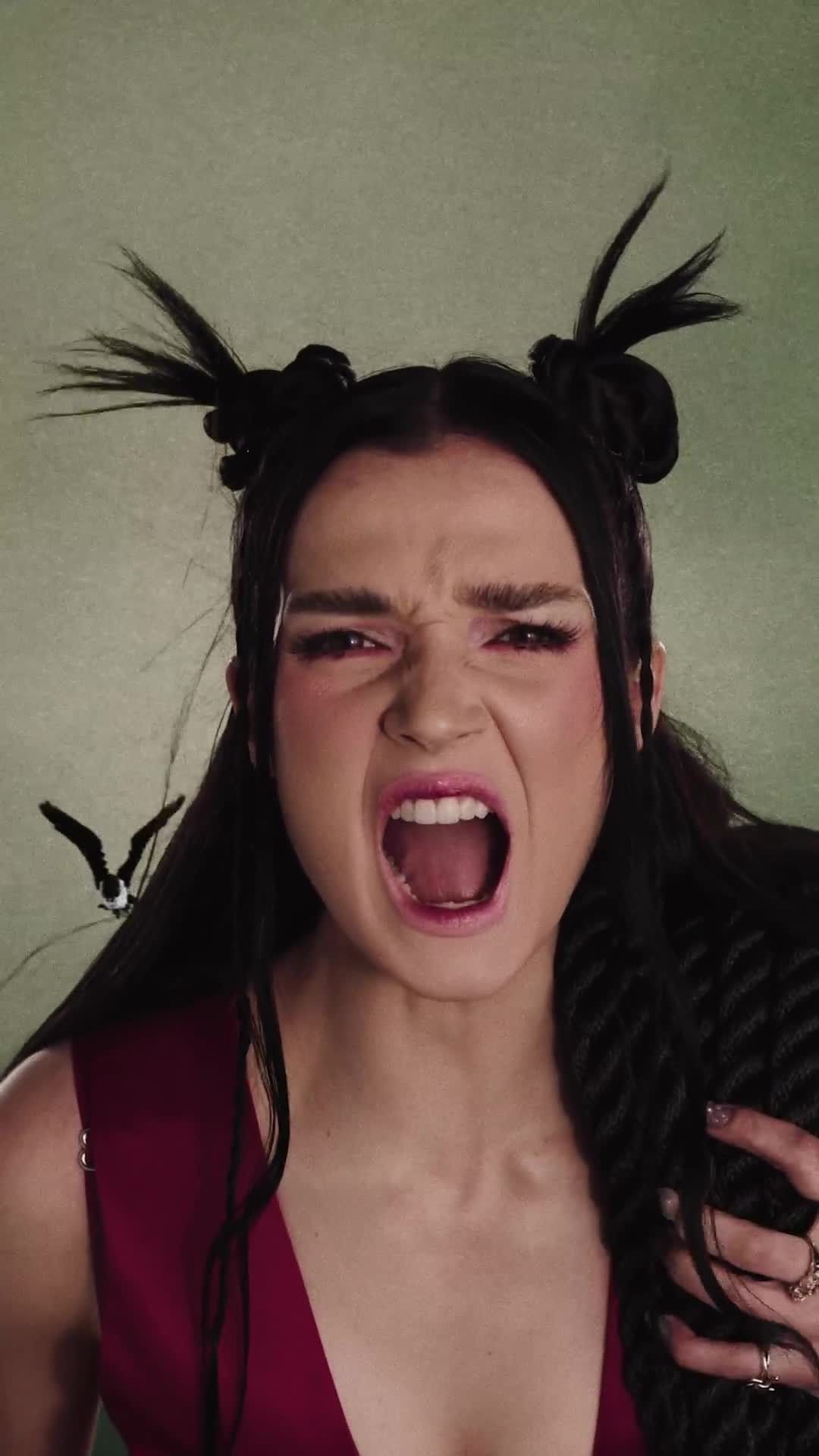 Portrait of an expressive adult woman with theatrical makeup, screaming with an open mouth, wearing a deep red top against a textured green backdrop.