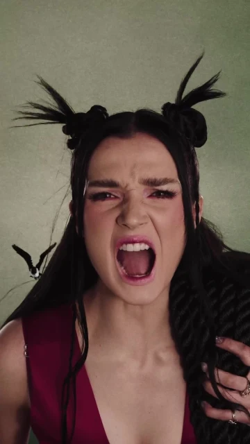 Portrait of an expressive adult woman with theatrical makeup, screaming with an open mouth, wearing a deep red top against a textured green backdrop.