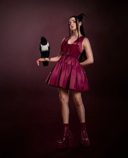 A woman in a burgundy dress poses in a studio with a magpie on her arm, creating a striking contrast against a dark backdrop.