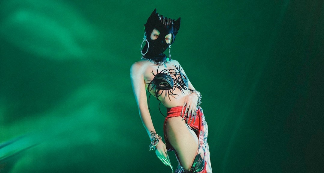 Person wearing a black cat mask, artistic bra, and red bottoms poses in front of a bright green background with artistic lighting.