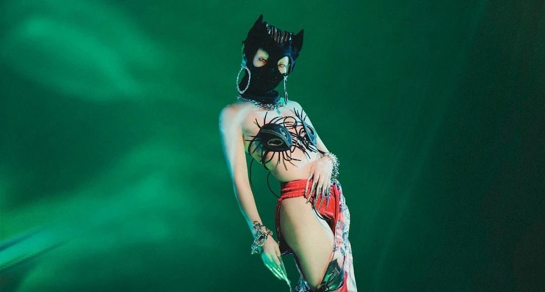 Person wearing a black cat mask, artistic bra, and red bottoms poses in front of a bright green background with artistic lighting.