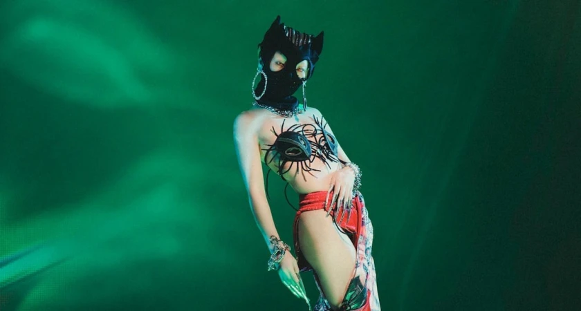 Person wearing a black cat mask, artistic bra, and red bottoms poses in front of a bright green background with artistic lighting.