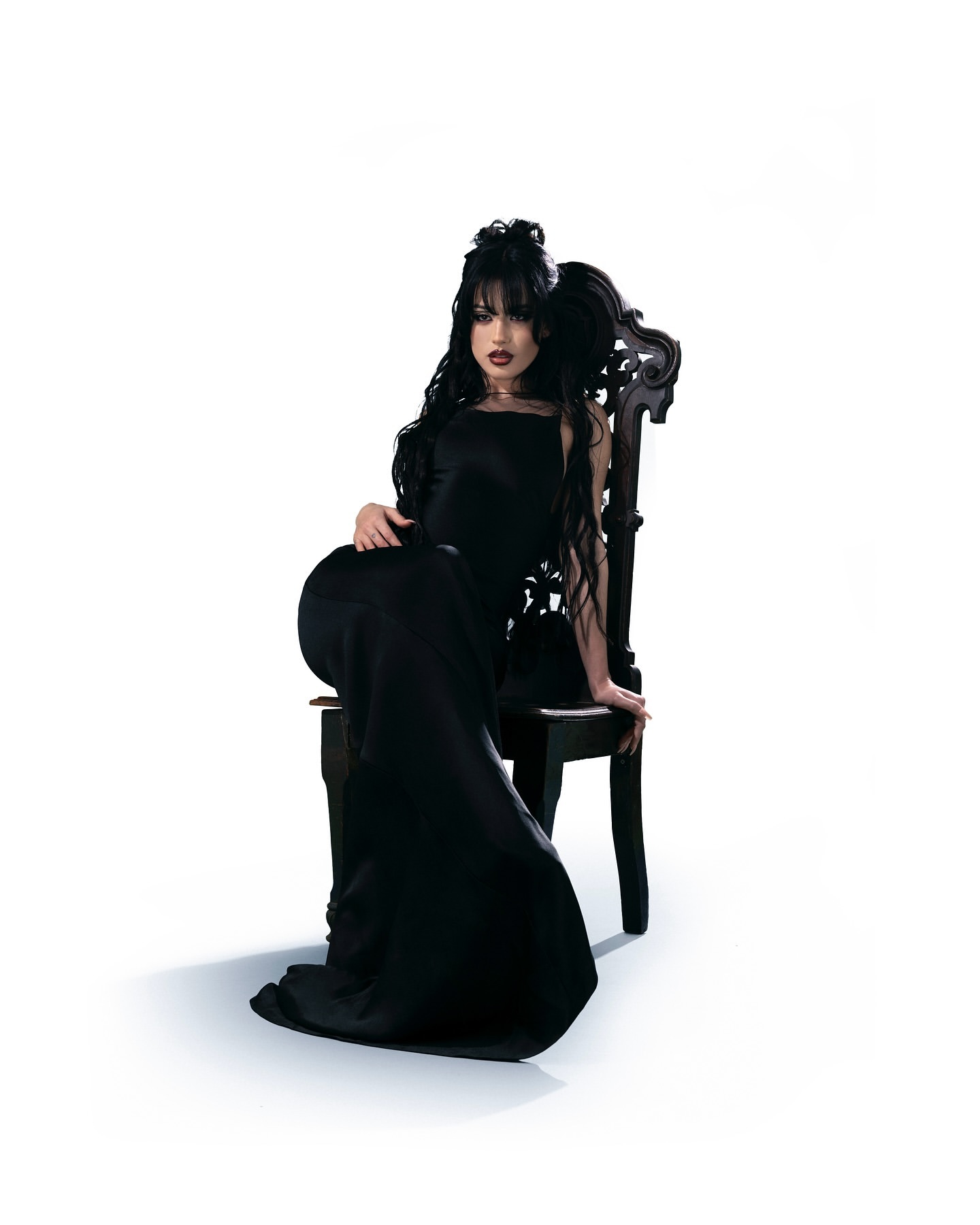 Woman in a long black dress seated on an ornate dark wooden chair against a white background.