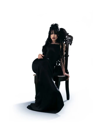 Woman in a long black dress seated on an ornate dark wooden chair against a white background.