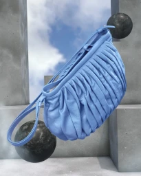 Blue leather handbag suspended against a surreal architectural concrete backdrop with dark spheres and sky