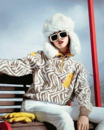 Model wearing a large white fluffy ushanka hat, square sunglasses, and zebra print fleece jacket seated outdoors.