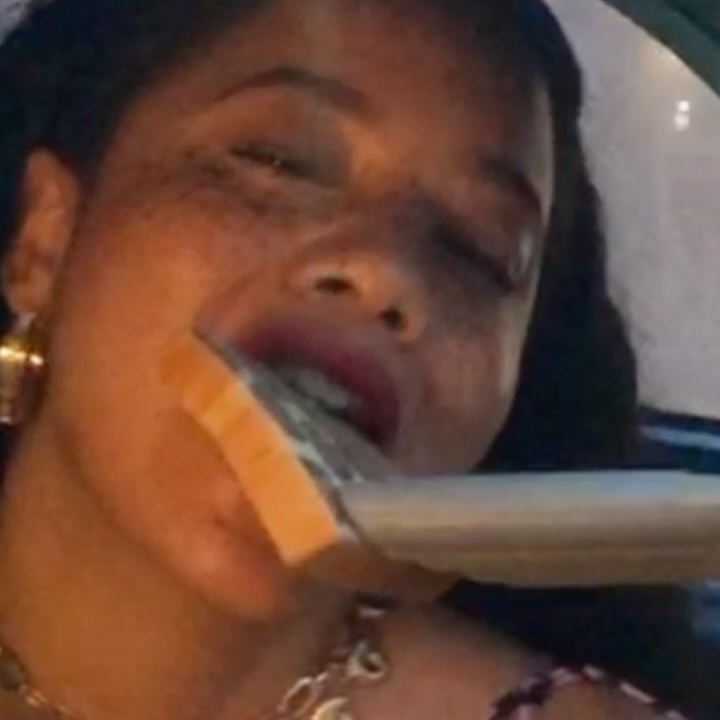 Close-up portrait of a woman with freckles holding currency in her mouth, wearing gold jewelry, under dim lighting.