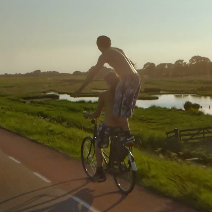 Two shirtless men on a bicycle on a paved path next to a grassy field with a body of water under a blue sky with light clouds.