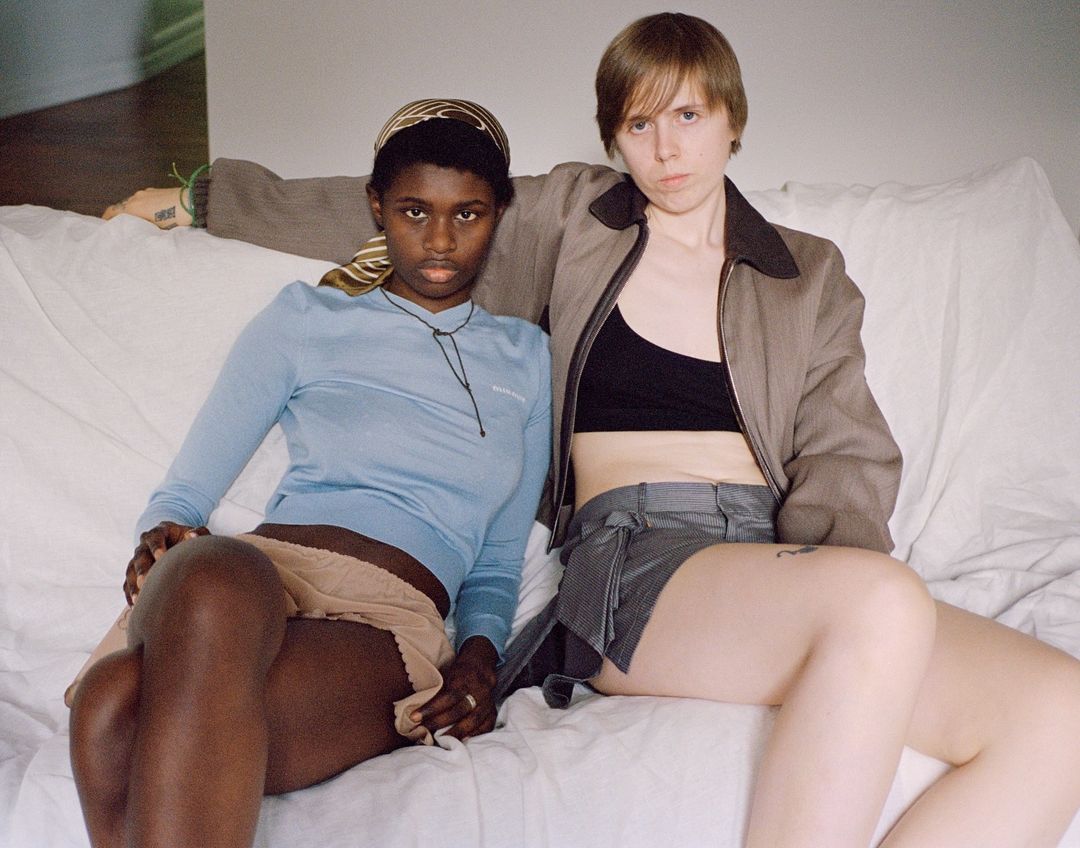 Two young adults seated indoors on a white sofa. One adult with a headscarf in a blue long-sleeved shirt and another with short hair.