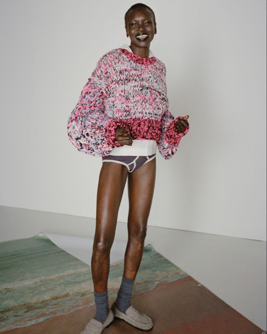 Woman in oversized knit sweater and socks poses on beach-themed paper in a studio with a white backdrop and soft, diffused lighting.