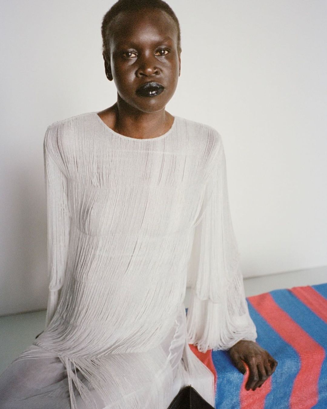 Dark-skinned model poses indoors wearing a white fringed dress, seated on a striped surface, portrait-style, in a fashion setting.