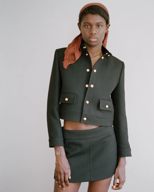 Woman in a dark green skirt suit with gold buttons and a reddish-brown headscarf posing in front of a plain light gray background.