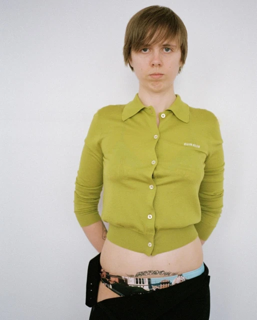 Portrait of a woman with short brown hair, wearing a lime-green cardigan, standing against a white wall in an indoor setting.