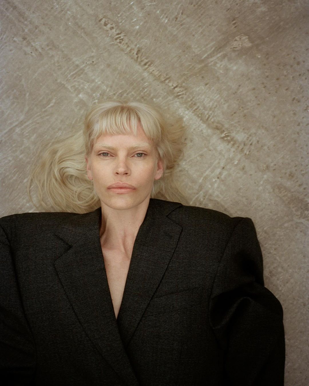 Blonde woman with bangs lying on a textured gray surface wearing a dark grey blazer in a close-up portrait, fashion photography.
