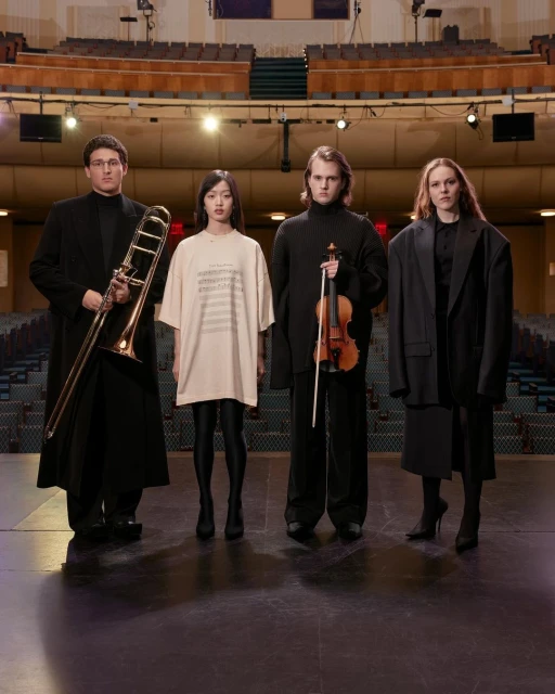 Four musicians stand in formal wear on a stage holding a trombone, violin; in the background are empty theater seats.