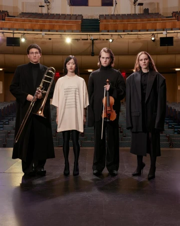 Four musicians stand in formal wear on a stage holding a trombone, violin; in the background are empty theater seats.