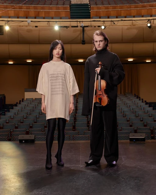 A casually dressed woman and a man in a turtleneck hold a violin on a theater stage with rows of empty seats and subdued lighting.