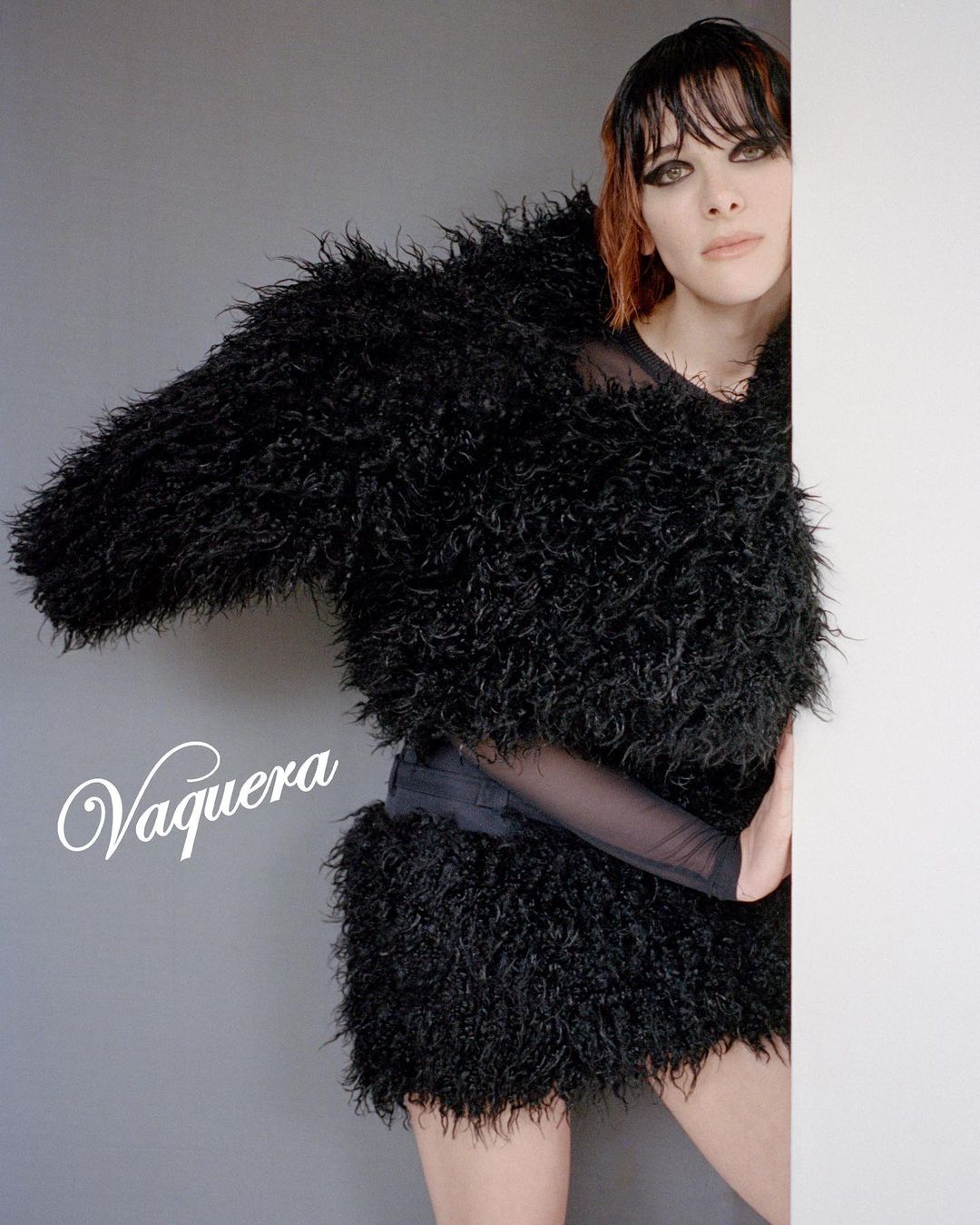 Model in textured black shaggy jacket peeking around a white wall corner against a grey studio background