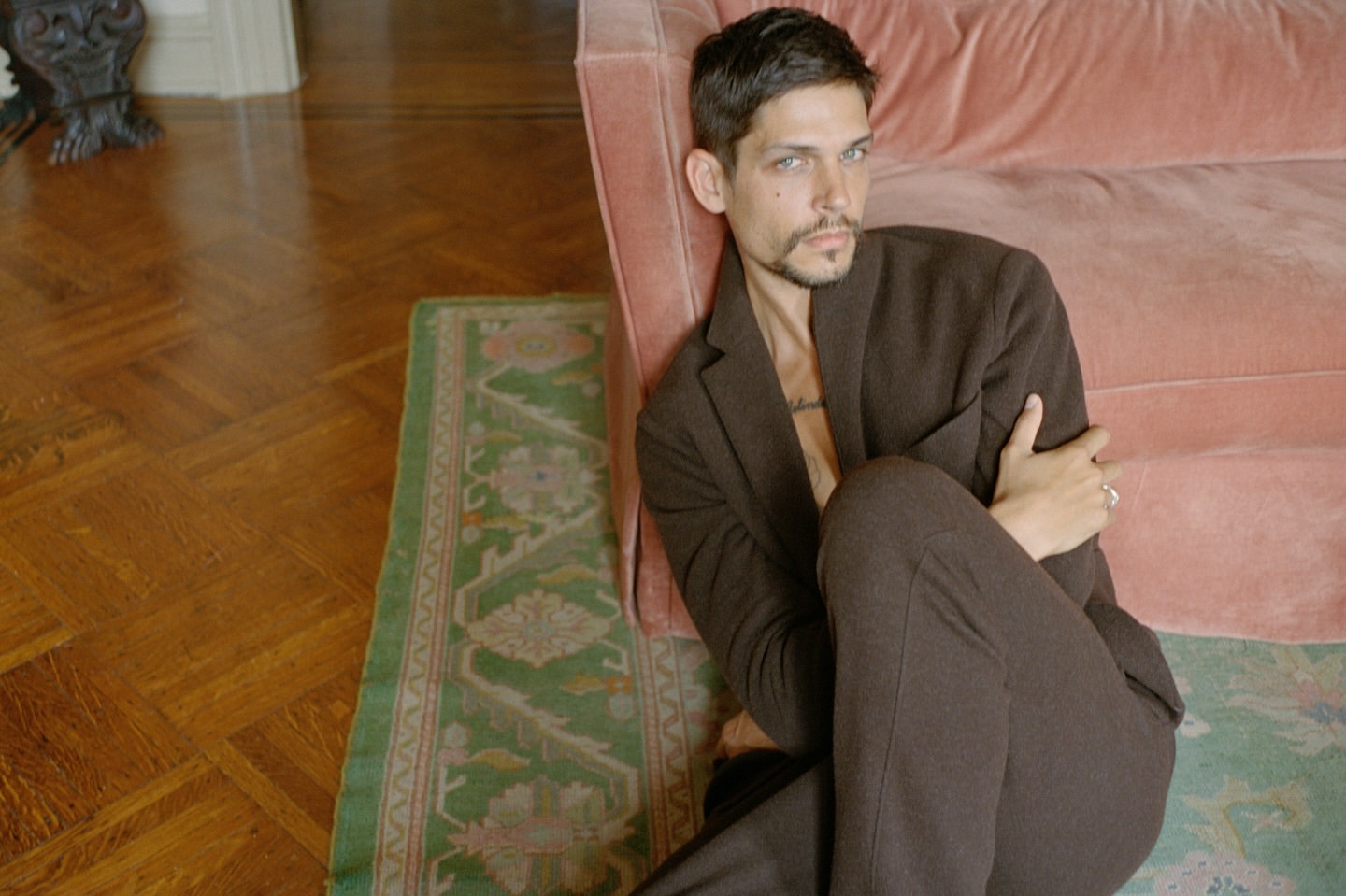 Adult male in a brown suit poses indoors near a pink couch and a green patterned rug on a parquet wood floor.