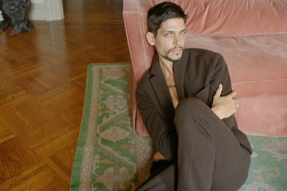 Adult male in a brown suit poses indoors near a pink couch and a green patterned rug on a parquet wood floor.