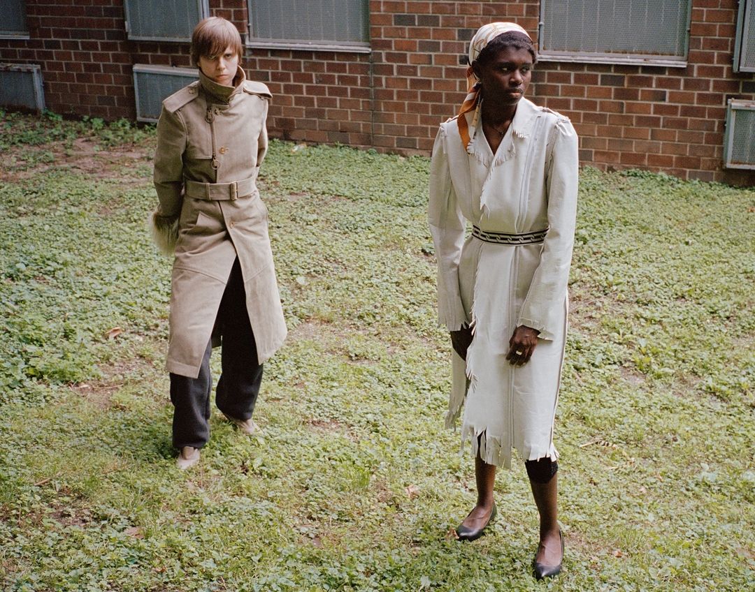 Two women in outdoor setting wearing neutral-toned coats, posing on a grassy field in front of a brick building with partially-opened windows.