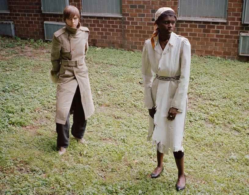 Two women in outdoor setting wearing neutral-toned coats, posing on a grassy field in front of a brick building with partially-opened windows.